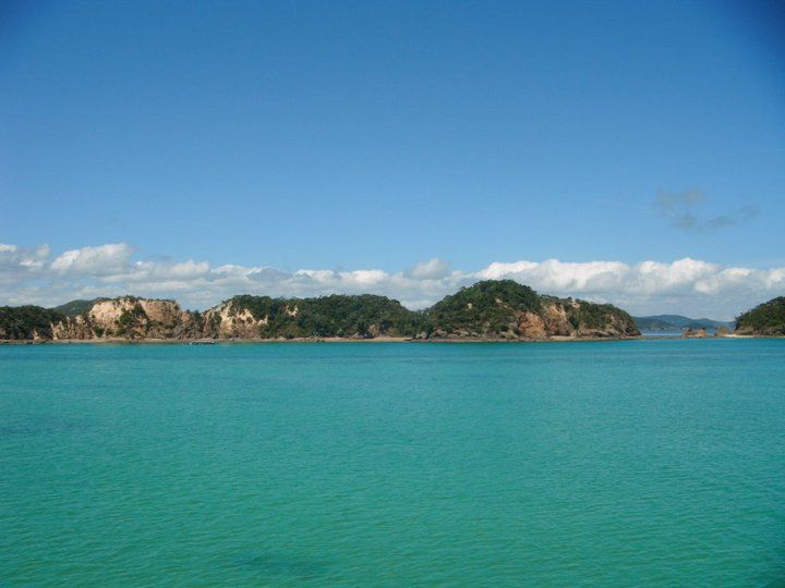 Stunning panoramic view of the Bay of Islands showing turquoise waters, lush green islands with rocky cliffs, and a clear blue sky with scattered white clouds