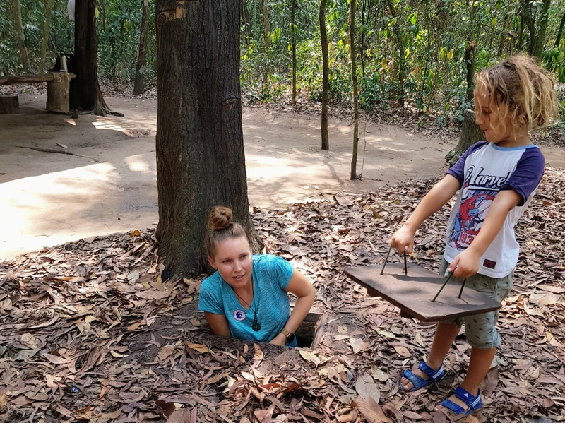 Family exploring the Cu Chi Tunnels with a woman emerging from a camouflaged tunnel entrance and a young boy holding a wooden cover with metal spikes