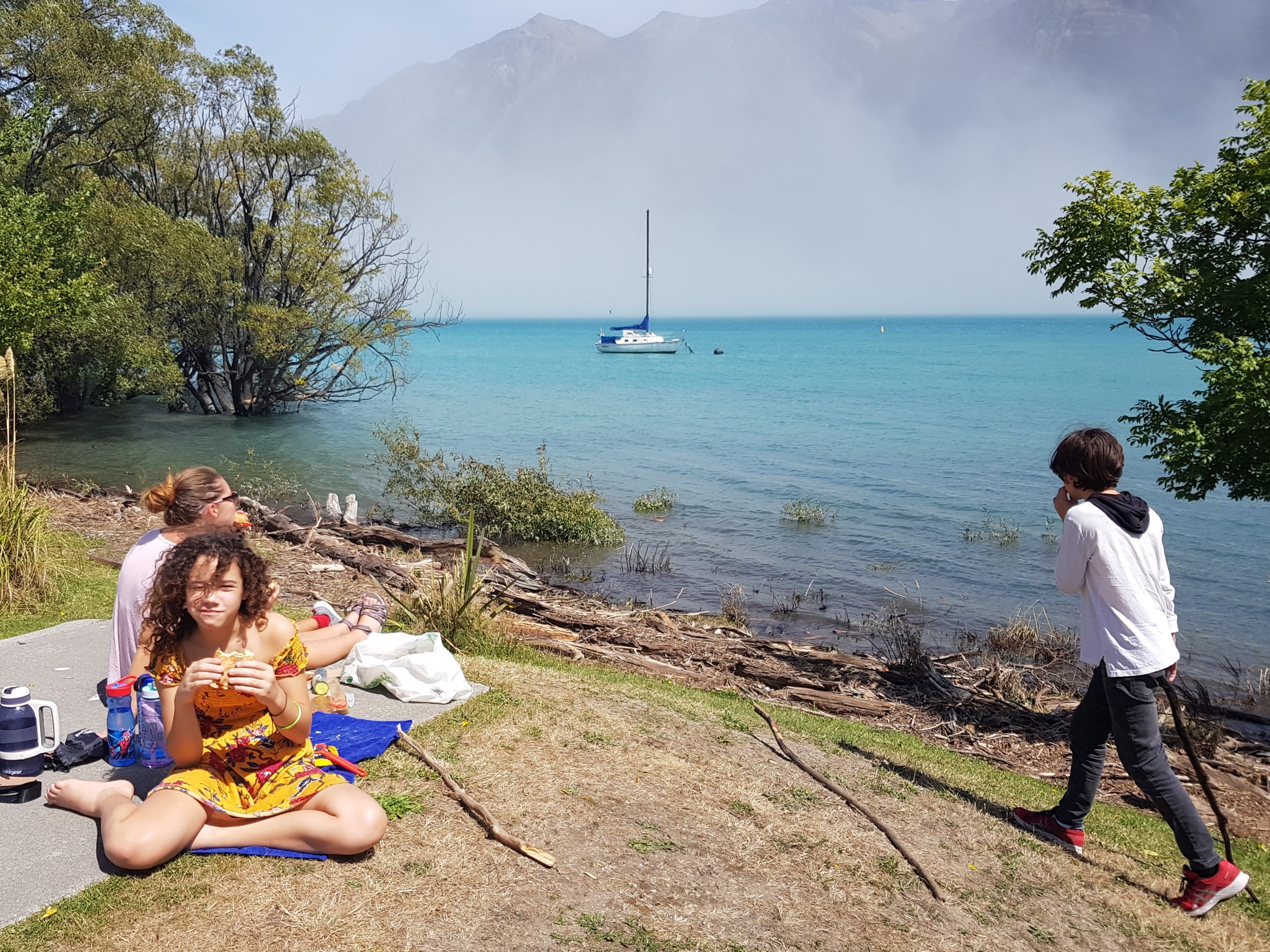 Family enjoying picnic lunch by turquoise lake in Glenorchy with sailboat and stunning mountain views in New Zealand