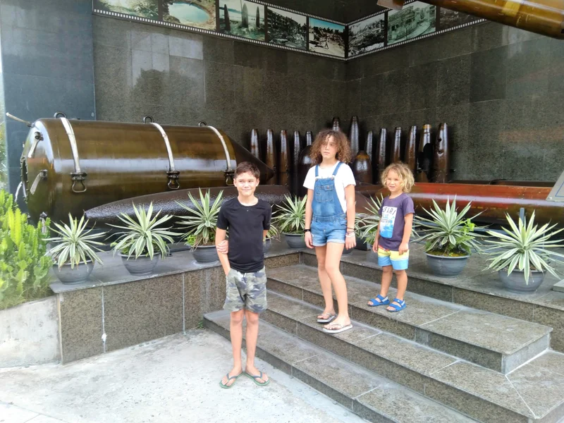 Three children standing on granite steps in front of war exhibits at the War Remnants Museum in Saigon, with historical artifacts and photographs displayed behind them